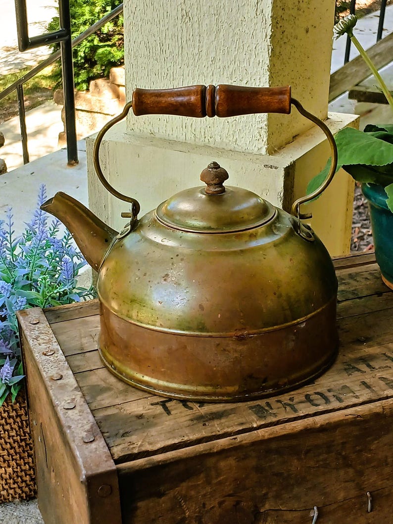 May include: Antique brass kettle with a wooden handle. The kettle has a rounded body, a spout, and a lid with a small knob. The handle is made of two pieces of wood joined together. The kettle sits on a wooden crate.