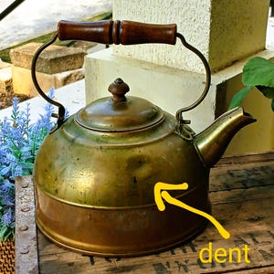 May include: Antique brass kettle with a wooden handle and a spout. The kettle has a round body with a lid and a handle that arches over the top. The kettle has a dent on the side.