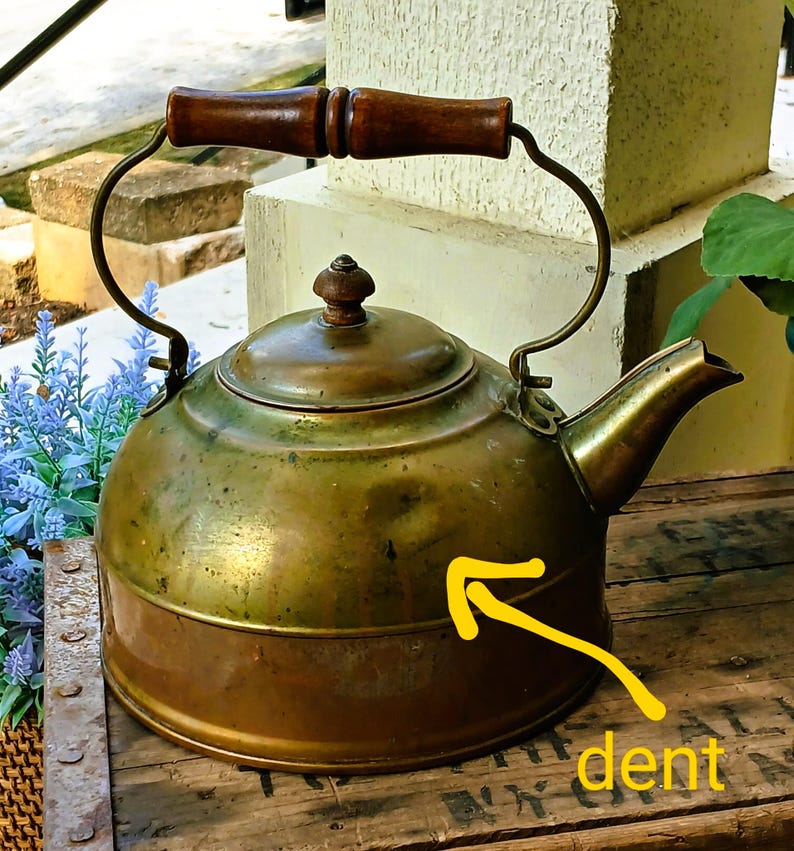 May include: Antique brass kettle with a wooden handle and a spout. The kettle has a round body with a lid and a handle that arches over the top. The kettle has a dent on the side.