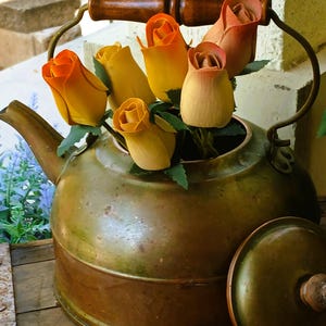 May include: An antique brass kettle with a wooden handle and lid, filled with yellow and orange wooden roses. The kettle sits on a wooden surface, with a glimpse of a garden in the background.