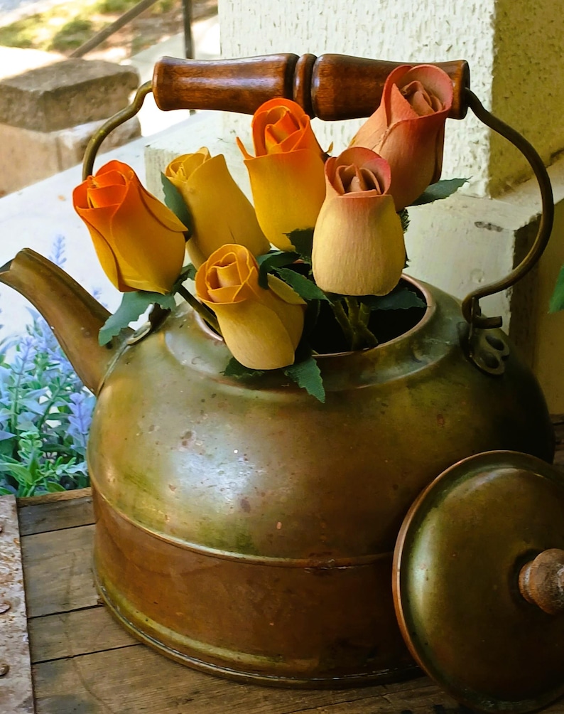 May include: An antique brass kettle with a wooden handle and lid, filled with yellow and orange wooden roses. The kettle sits on a wooden surface, with a glimpse of a garden in the background.