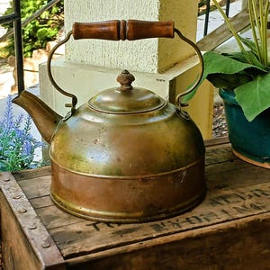 May include: A vintage copper kettle with a wooden handle and brass accents. The kettle sits on a wooden crate, with a green plant in a blue pot and lavender flowers in the background. The kettle has a round body and a spout.