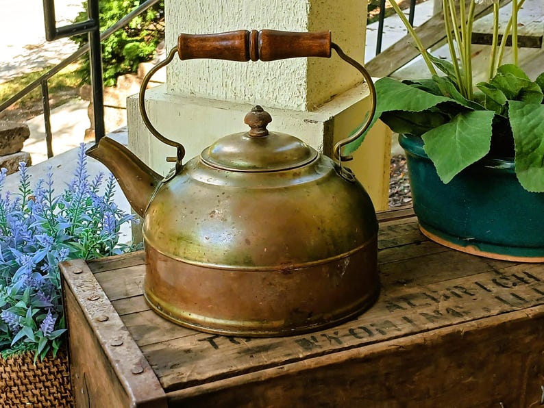 May include: A vintage copper kettle with a wooden handle and brass accents. The kettle sits on a wooden crate, with a green plant in a blue pot and lavender flowers in the background. The kettle has a round body and a spout.