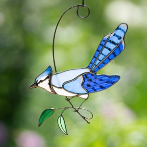 May include: A stained glass blue jay with a white chest and black eyes perched on a green branch. The bird is hanging from a wire loop.