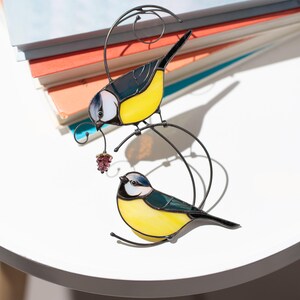 Stained glass blue tit suncatcher placed on a table surrounded by colorful glass sheets, handmade bird window decor with yellow and blue feathers, nature-inspired decoration.