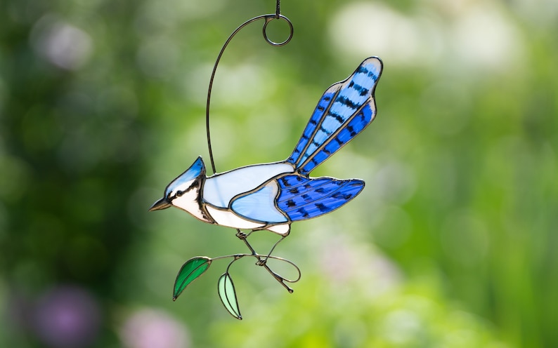 May include: A stained glass blue jay with a white chest and black eyes perched on a green branch. The bird is hanging from a wire loop.