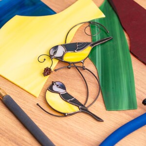 Stained glass blue tit suncatcher placed on a table surrounded by vibrant colored glass sheets, showcasing yellow and blue bird design, handmade nature-inspired decor.