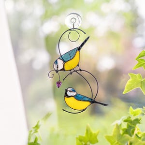 Stained glass blue tit suncatcher held in hand near a window, vibrant yellow and blue bird design with a purple flower, decorative handmade window hanging.