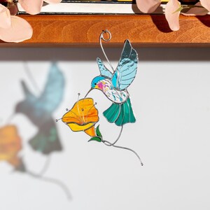stained glass hummingbird suncatches hanging from a wooden shelf casting colorful bird shadow on the wall. Handmade stained glass bird window decoration inspired by hummingbird and bright spring flower