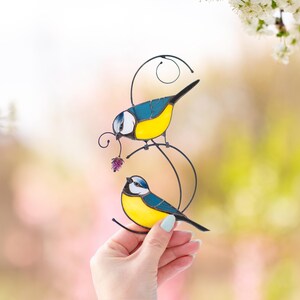 Stained glass blue tit suncatcher held in hand against a floral soft background, with bright sunlight filtering through, handmade bird window hanging with yellow and blue feathers.