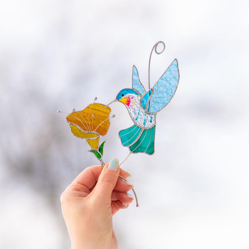 Hand holding a stained glass hummingbird suncatcher with blue wings and orange flower against a soft neutral background. Handmade stained glass bird window hanging designed as a colorful nature inspired decor piece.
