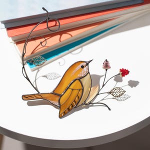 Stained glass carolina wren bird decor displayed beside stacked books, handcrafted bird suncatcher with floral wire branch.