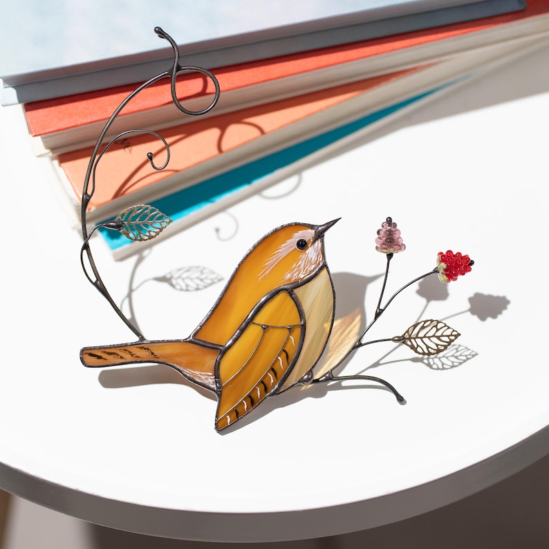 Stained glass carolina wren bird decor displayed beside stacked books, handcrafted bird suncatcher with floral wire branch.