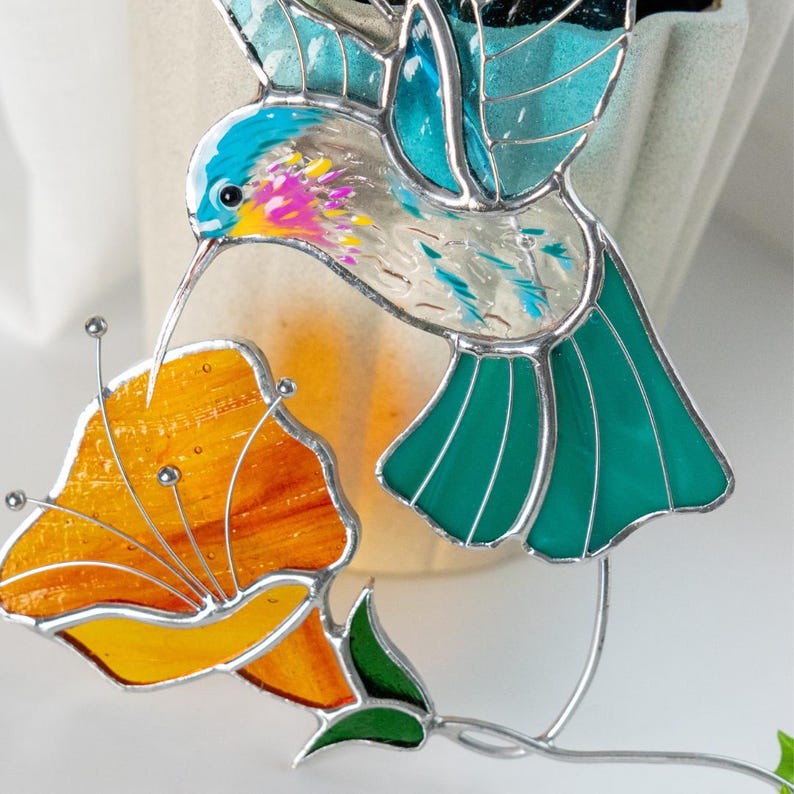 Close view of a handmade stained glass hummingbird suncatcher with textured blue wings and bright orange flower. Decorative stained glass bird window hanging inspired by hummingbirds and blooming garden flowers.