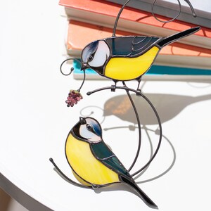 Stained glass blue tit suncatcher placed on a table surrounded by colorful glass sheets, handmade bird window decor with yellow and blue feathers, nature-inspired decoration.
