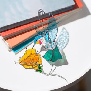Stained glass hummingbird suncatcher displayed with colored glass sheets. Handmade stained glass bird window hanging with turquoise wings and orange flower showing artisan glass materials.