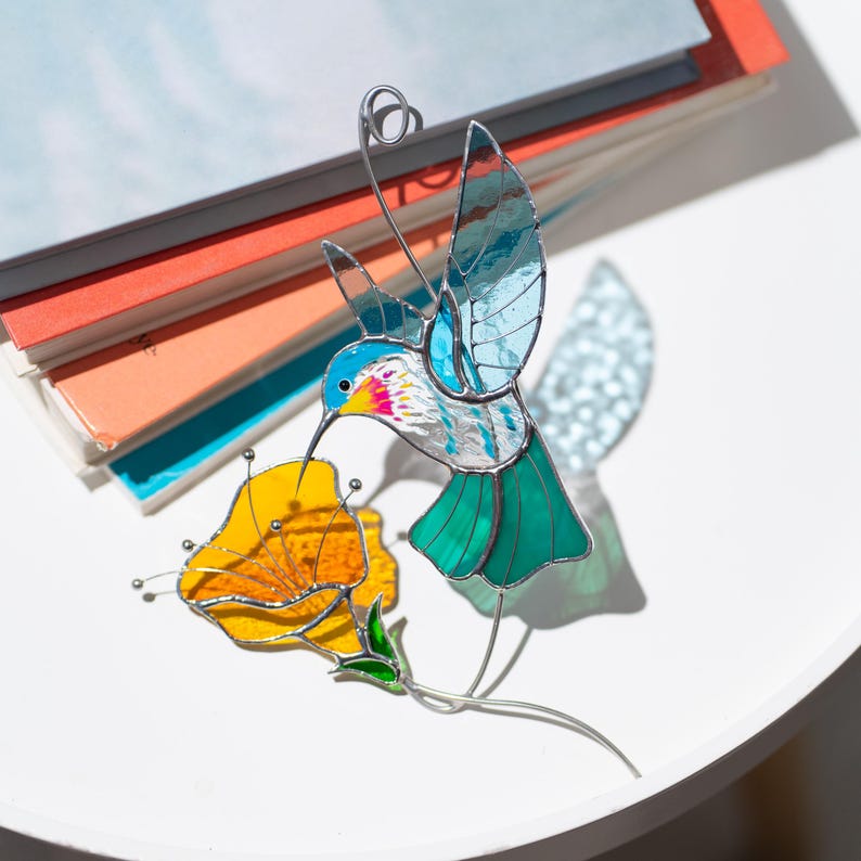 Stained glass hummingbird suncatcher displayed with colored glass sheets. Handmade stained glass bird window hanging with turquoise wings and orange flower showing artisan glass materials.