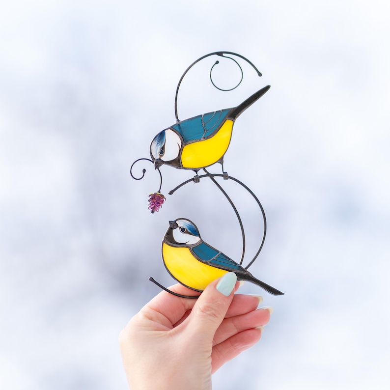 Hand holding stained glass blue tit suncatcher against soft natural light background, handmade window hanging decoration.