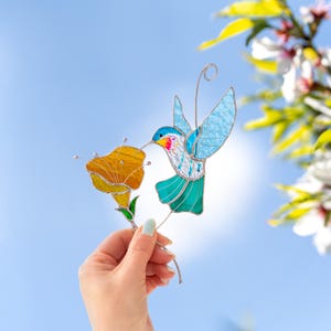 Hand holding a stained glass hummingbird suncatcher with turquoise wings and orange flower against a blue sky background. Handmade stained glass bird window decoration designed to catch sunlight and brighten a room.