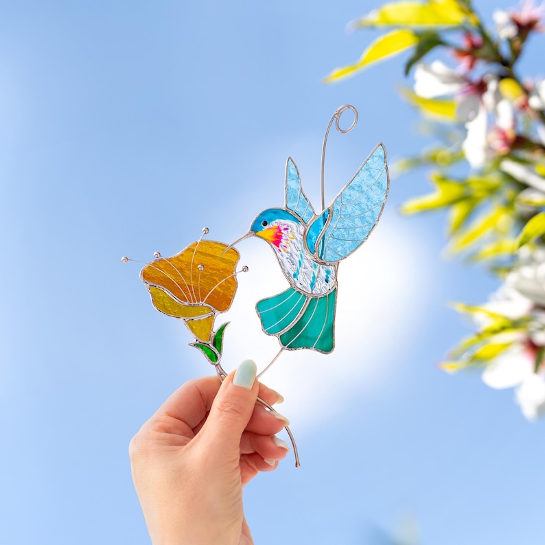 Hand holding a stained glass hummingbird suncatcher with turquoise wings and orange flower against a blue sky background. Handmade stained glass bird window decoration designed to catch sunlight and brighten a room.