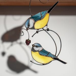 Stained glass blue tit suncatcher casting a shadow on a white surface, bright yellow and blue bird design, hanging with fine metalwork details.