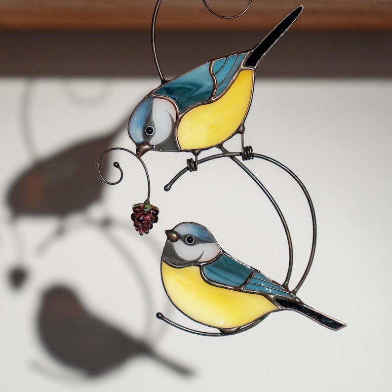 Stained glass blue tit suncatcher casting a shadow on a white surface, bright yellow and blue bird design, hanging with fine metalwork details.