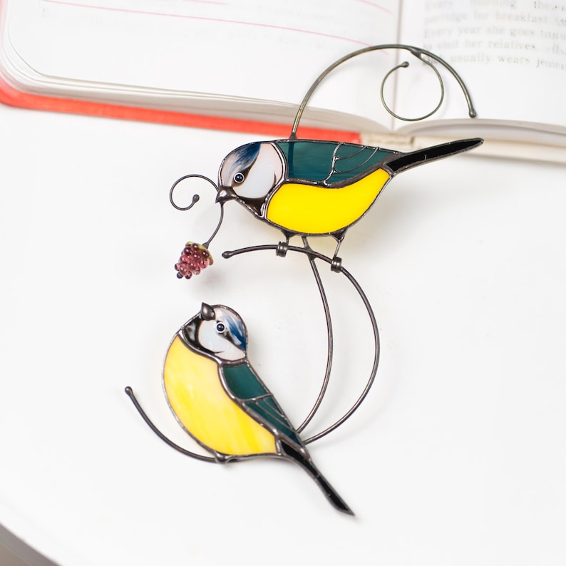 Stained glass blue tit suncatcher placed on a table surrounded by colorful glass sheets, handmade bird window decor with yellow and blue feathers, nature-inspired decoration.