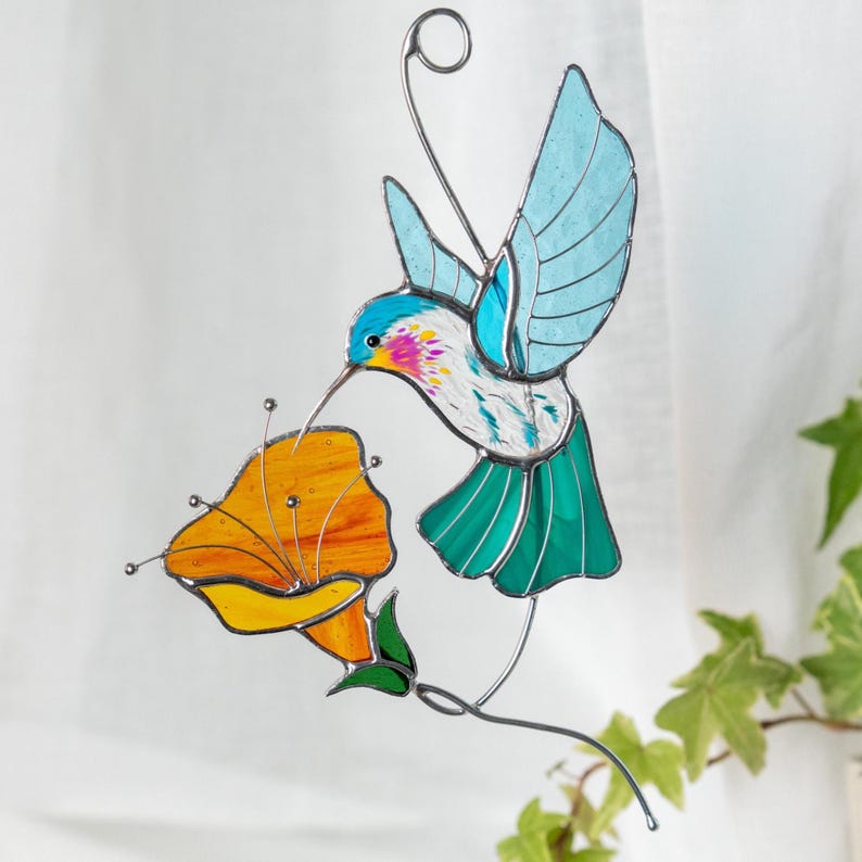 Stained glass hummingbird suncatcher with turquoise wings hovering above an orange flower, handmade stained glass bird window hanging displayed against a light background. Nature inspired stained glass decor for window or garden.