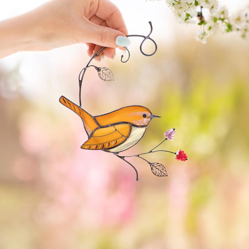 Hand holding handmade stained glass carolina wren suncatcher with floral branch, cheerful window hanging decor in sunlight.