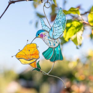 Handmade stained glass hummingbird suncatcher hanging outdoors on a tree branch with sunlight passing through blue wings and an orange poppy flower. Artistic stained glass bird window decoration inspired by garden nature and spring colors.