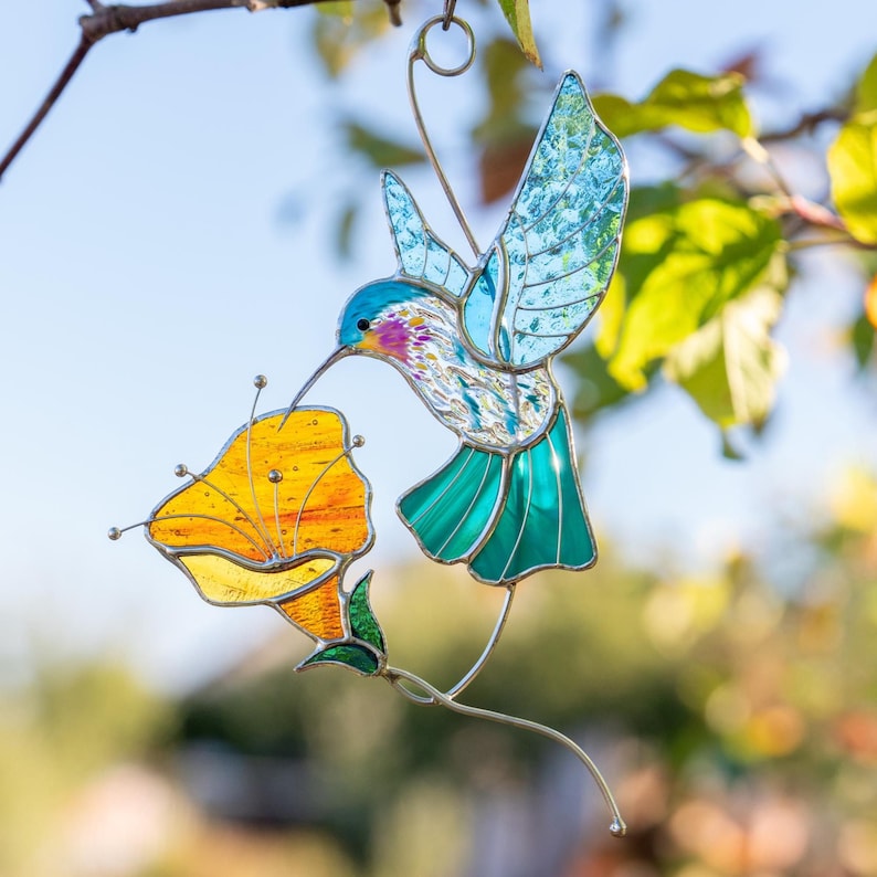 Handmade stained glass hummingbird suncatcher hanging outdoors on a tree branch with sunlight passing through blue wings and an orange poppy flower. Artistic stained glass bird window decoration inspired by garden nature and spring colors.