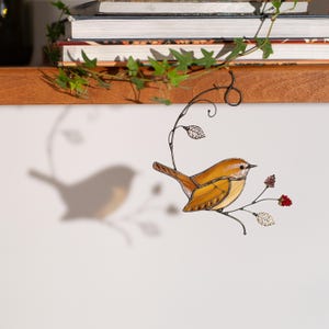 Handmade stained glass Carolina Wren hanging near books and ivy, bird shadow reflected on wall with floral branch details.