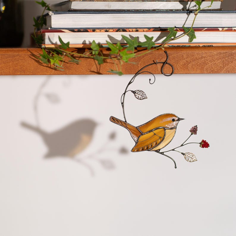 Handmade stained glass Carolina Wren hanging near books and ivy, bird shadow reflected on wall with floral branch details.