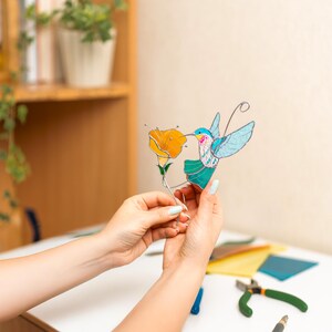 Hands holding a stained glass hummingbird suncatcher near a workspace with glass tools and materials. Handmade stained glass bird decoration featuring turquoise wings and an poppy flower inspired by nature.