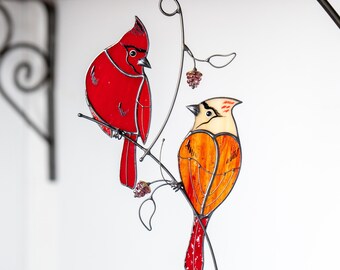 May include: A stained glass cardinal and a stained glass bird with orange and red plumage perched on a black metal branch with a berry.
