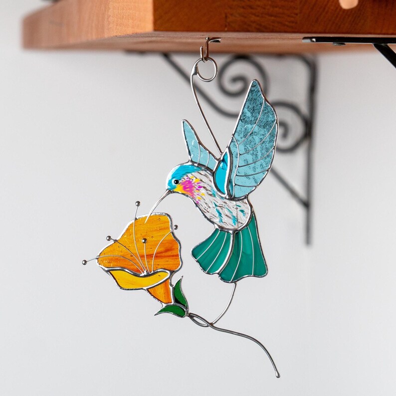 Stained glass hummingbird suncatcher hanging on a decorative metal bracket outdoors. Handmade stained glass bird window hanging with blue wings and orange flower inspired by nature and garden decor.