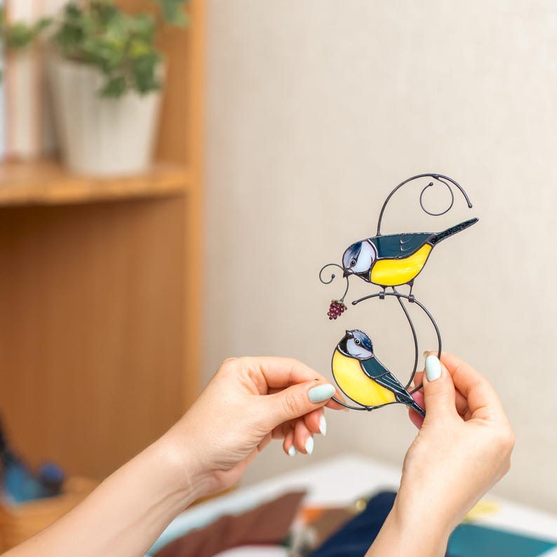 Stained glass blue tit suncatcher held by hands with a bright, blurred background, handmade bird window hanging with vivid yellow and blue colors, artistic bird decor.