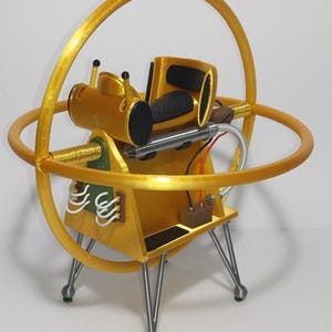 Op de afbeelding: Een goudkleurig, futuristisch uitziend apparaat met een cirkelvormig frame en een centraal compartiment. Het heeft een cilindrisch onderdeel, zwarte vulling en zilveren poten. Het ontwerp bevat mechanische en elektronische elementen, wat wijst op een sciencefictionthema.