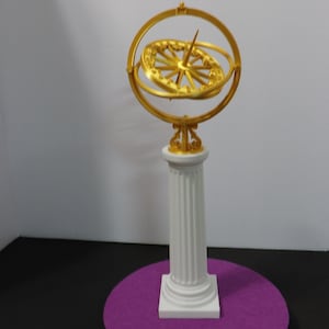 Puede incluir: Una esfera armilar dorada, modelo de objetos celestes, sobre una columna blanca con base cuadrada. La columna descansa sobre una base circular morada, sobre una superficie negra. La esfera tiene un dial central y anillos.