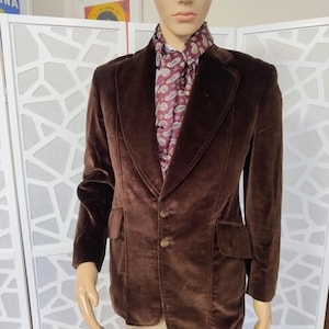 Puede incluir: Un blazer de terciopelo marrón oscuro con solapa de muesca, dos botones frontales y dos bolsillos con solapa. El blazer se lleva sobre una bufanda granate con un patrón de cachemira en blanco y azul claro. La prenda se exhibe en un maniquí.