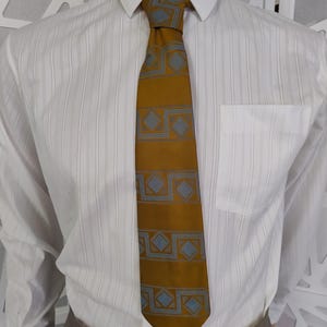 Puede incluir: Una corbata color mostaza con un patrón geométrico repetitivo en azul claro. La corbata se lleva con una camisa blanca con rayas verticales y un bolsillo. La camisa está abotonada en el cuello.