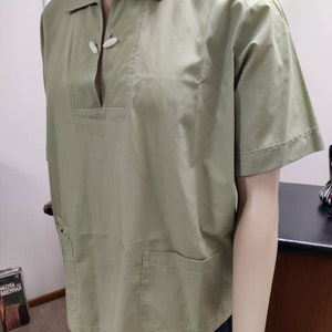 May include: A light green short-sleeved shirt with a collar and two pockets. The shirt has a unique button closure at the neck.