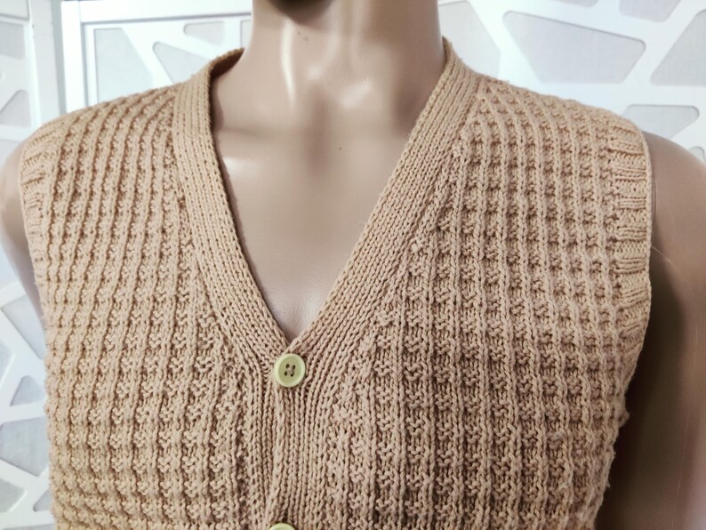 May include: A sleeveless knitted vest with a V-neck and button closure. The vest is a light brown color and has a waffle knit texture.