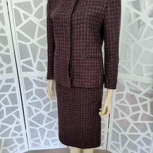 May include: A two-piece burgundy suit with a textured pattern. The suit includes a long-sleeved jacket with a single button closure and a matching knee-length skirt. The suit is displayed on a mannequin.