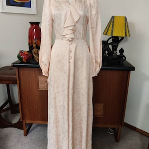 Peut inclure: Une longue robe en satin rose pâle avec un col à volants et un motif floral. La robe a des manches longues et un devant boutonné.