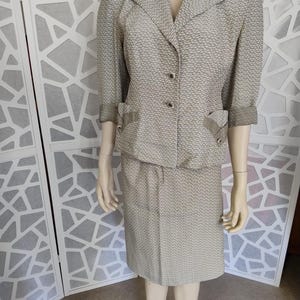 May include: A two-piece vintage suit in a neutral color palette. The jacket has a notched collar, three buttons, and two front pockets. The skirt is knee-length. The suit is made of a textured fabric with a subtle pattern.