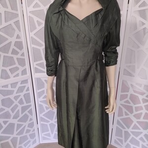 Puede incluir: Vestido vintage verde oliva con cuello ancho y mangas tres cuartos. El vestido presenta un corpiño entallado y una falda plisada, creando una silueta clásica. La tela tiene un brillo sutil, lo que aumenta su apariencia elegante.