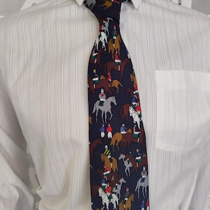 Puede incluir: Corbata azul marino con un patrón repetido de jinetes a caballo. Los jinetes visten atuendos coloridos, y los caballos son de varios tonos de marrón y gris. La corbata se lleva sobre una camisa blanca.