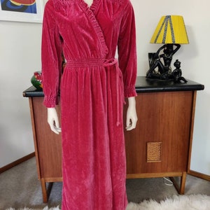 May include: A long pink velvet robe with a ruffled collar and a tie belt. The robe has long sleeves and a gathered waist.