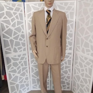 以下が含まれることがあります： マネキンに着せられたベージュのスーツ。2つボタンのジャケットとマッチするズボンで構成されています。白い襟付きシャツ、柄物のネクタイ、茶色のツイードキャップを合わせています。背景には白い折りたたみ式のスクリーンと額入りのポスターがあります。